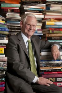 News of Doug Otto wearing a suit and tie and seated in front of huge stacks of books